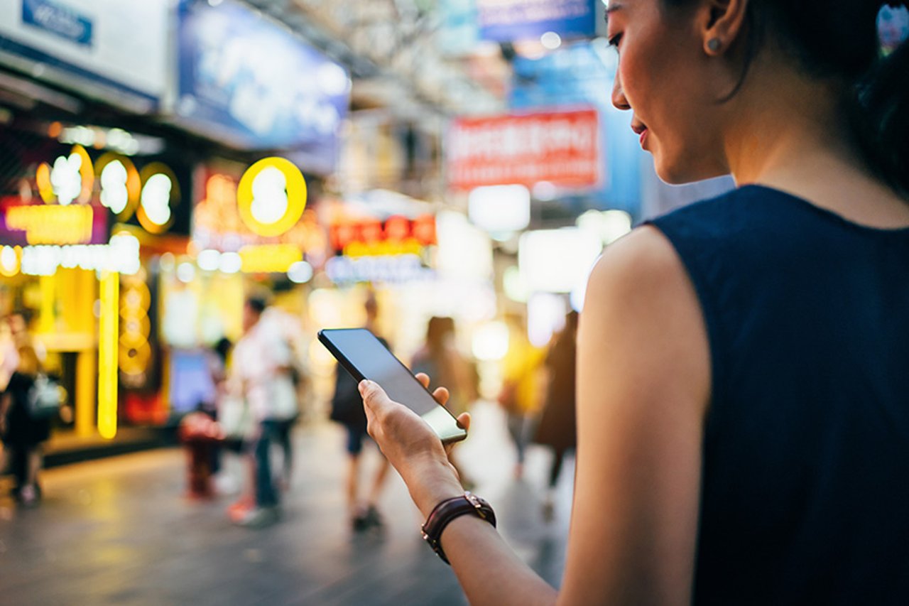 Woman using smartphone in busy city street, against colourful neon commercial sign and city buildings